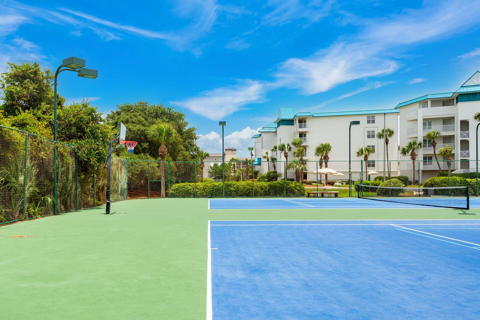 Un court de tennis avec un filet, entouré d’une clôture verte, et un bâtiment avec des balcons en arrière-plan.