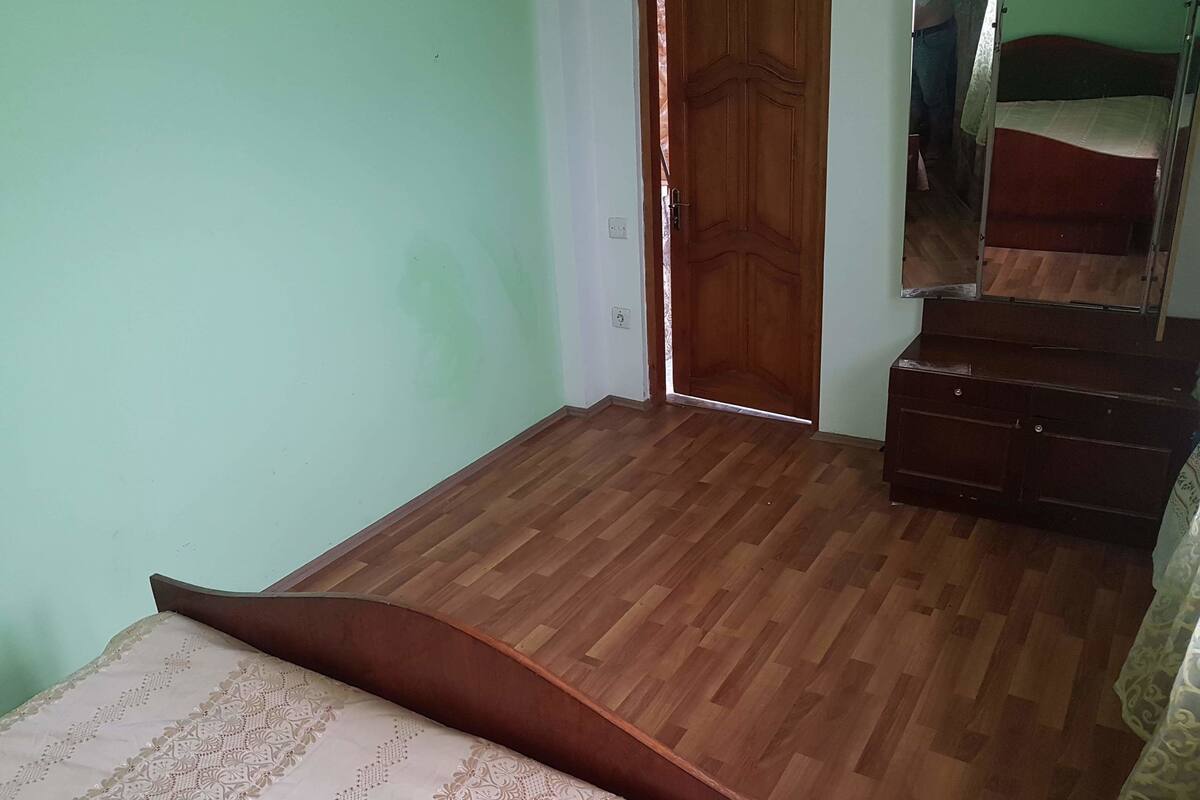 A bedroom with a wooden floor, a bed with a patterned bedspread, a wooden wardrobe, and a mirror on the wall.