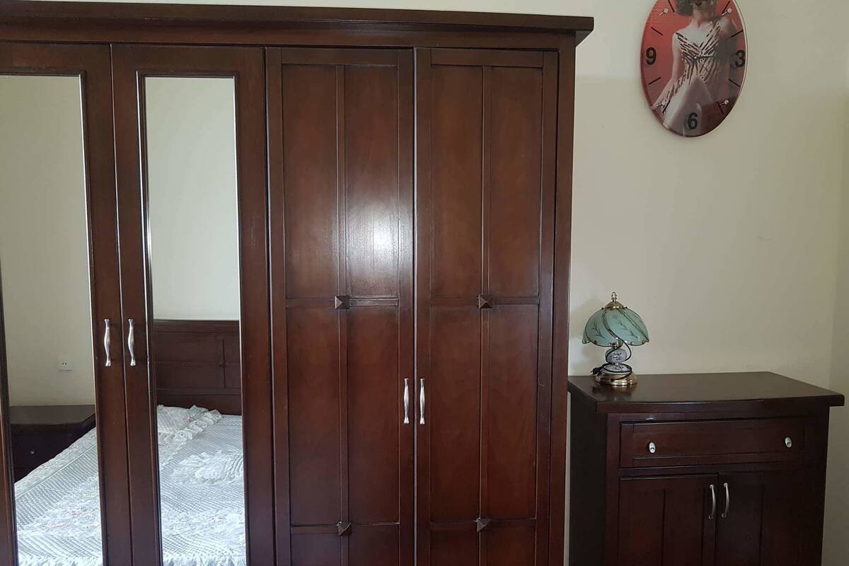 A bedroom with a wooden wardrobe, a bed with a white bedspread, and a wall clock.