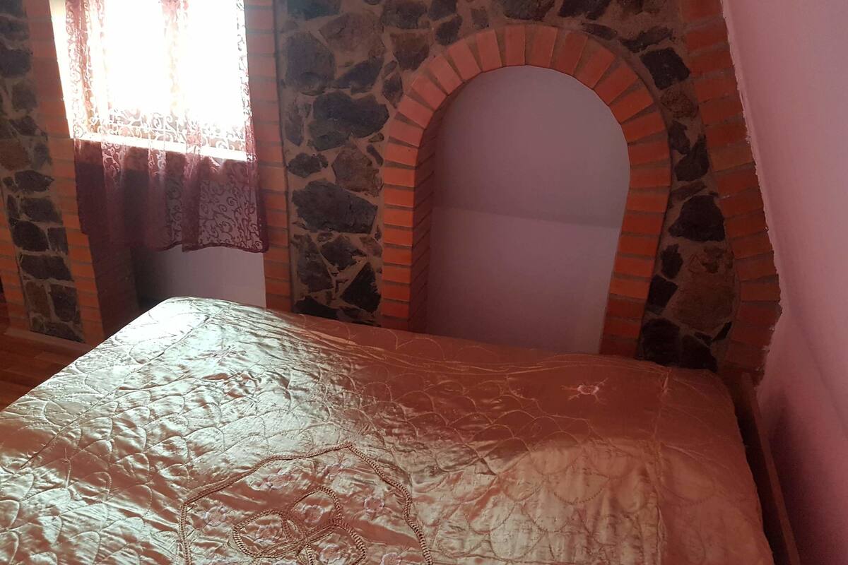 A room with a stone wall, a brick arch, a bed with a patterned bedspread, and a window with curtains.