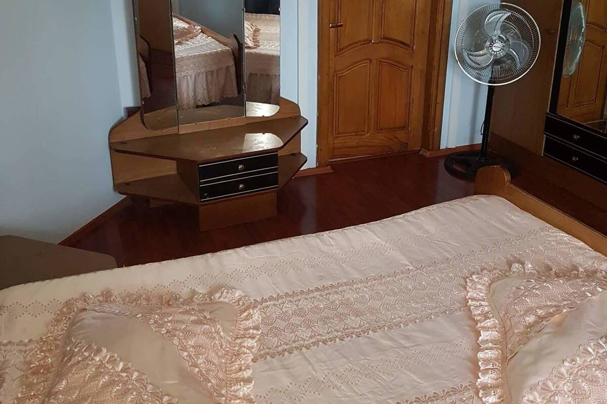 A bedroom with a bed, a wooden wardrobe, a mirror, and a standing fan.