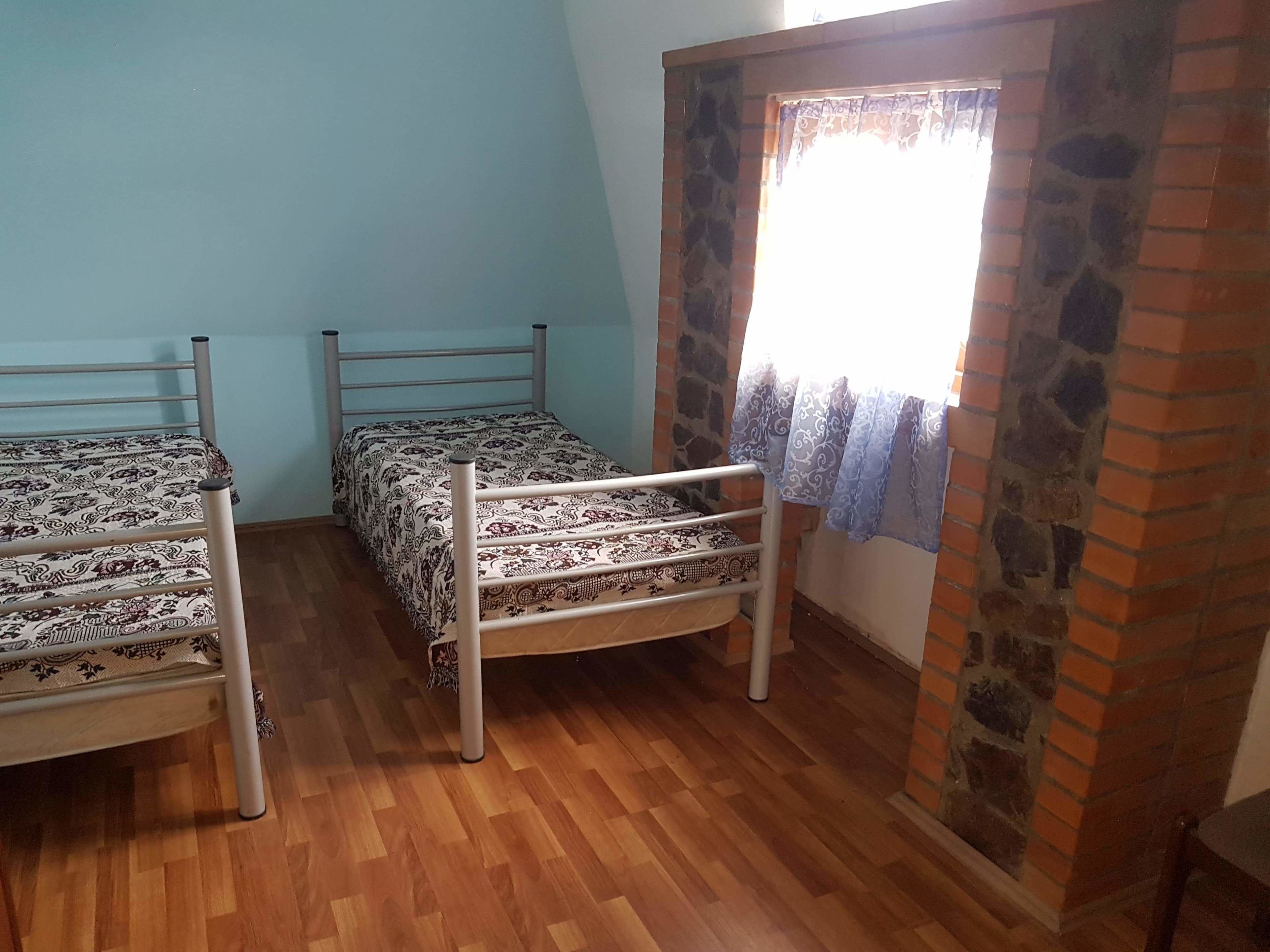 A room with two metal bunk beds with patterned bedding, a wooden floor, and a stone wall with a window covered by a patterned curtain.