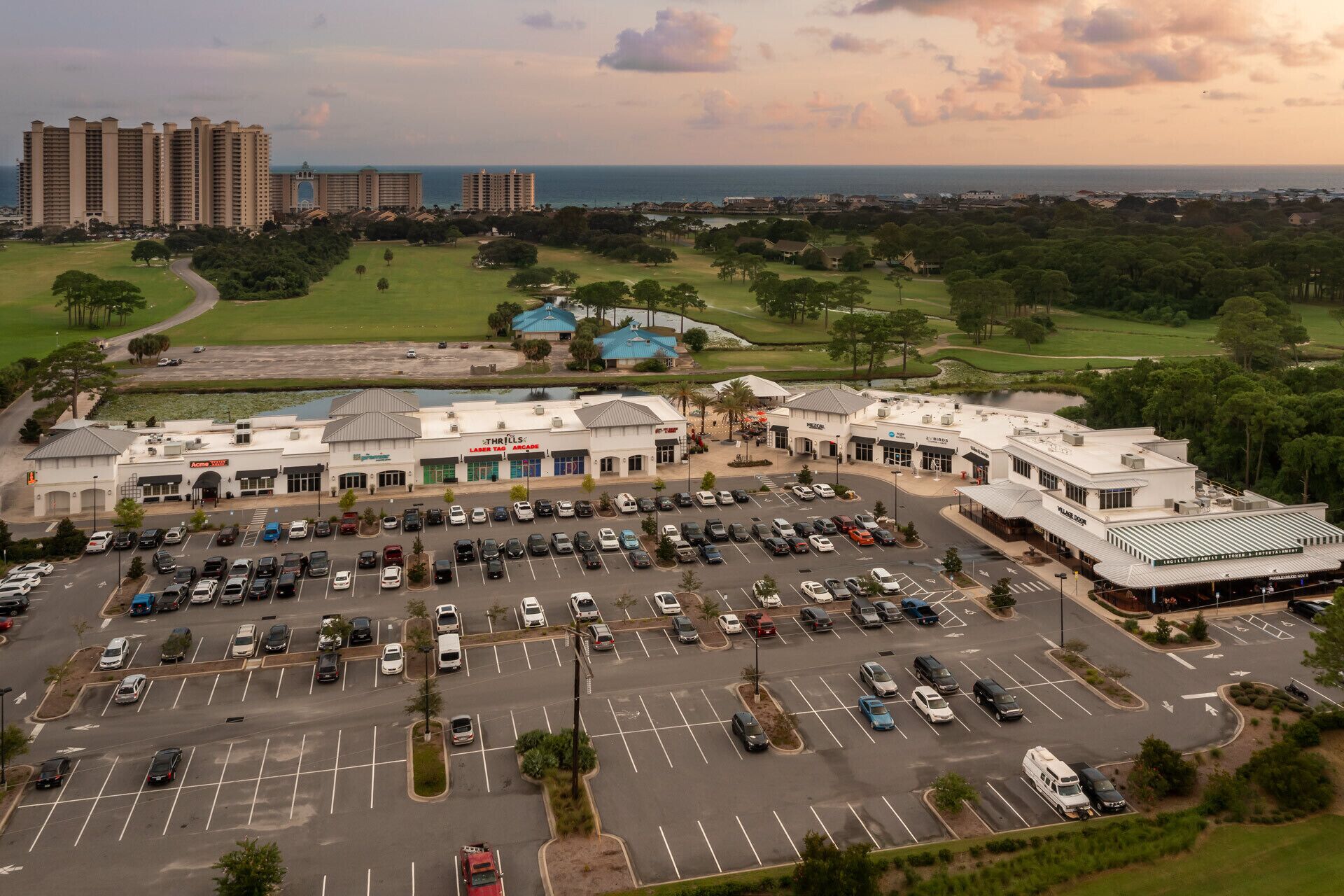 A large parking lot with multiple rows of cars, a shopping center with various stores, and a view of the ocean and residential buildings in the background.