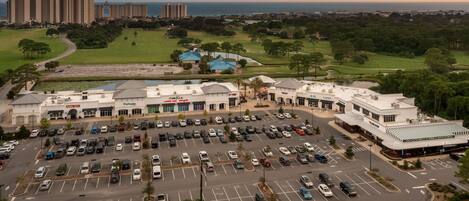 A large parking lot with multiple rows of cars, a shopping center with various stores, and a view of the ocean and residential buildings in the background.