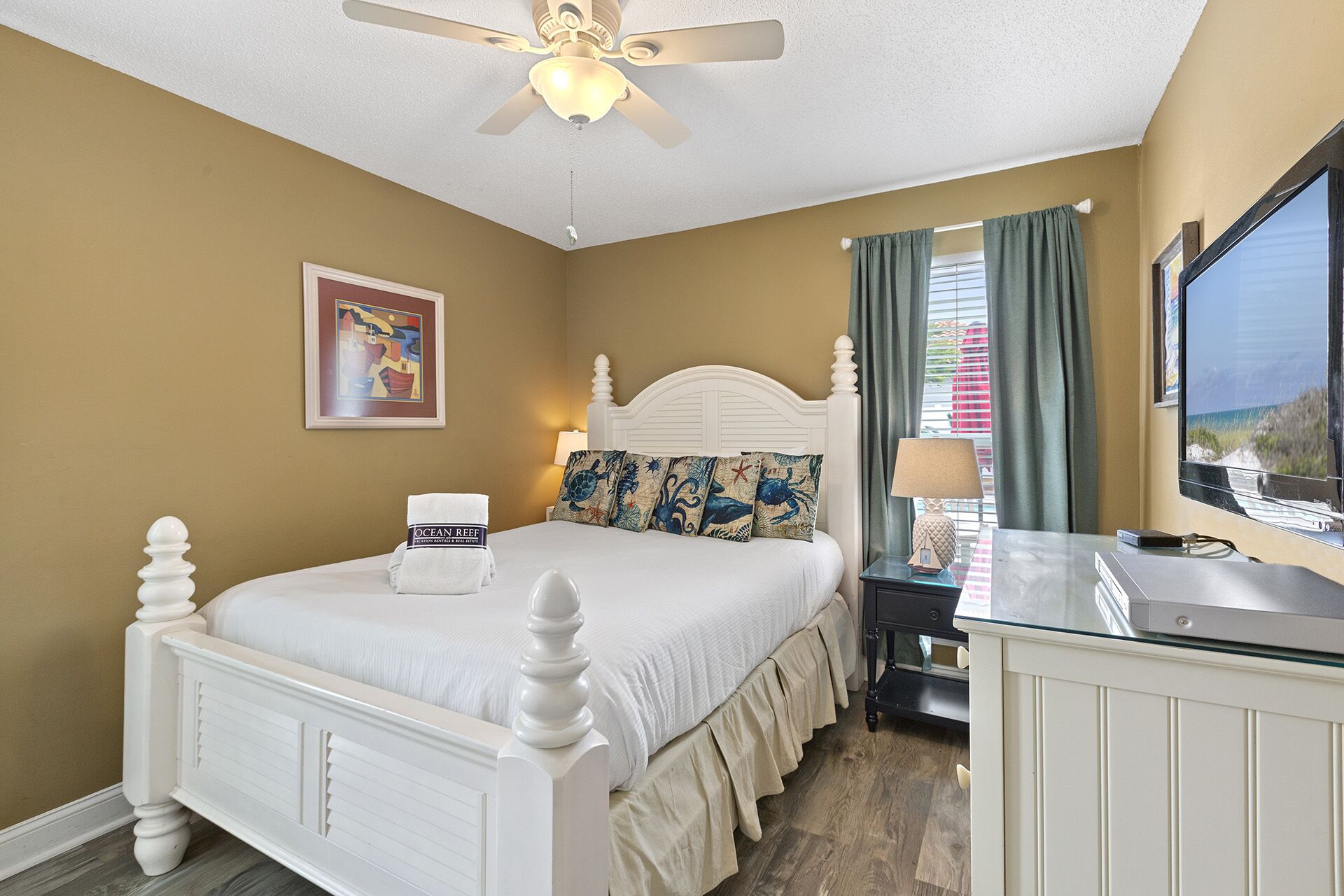 A hotel room with a bed, a desk, a ceiling fan, a window with curtains, and a painting on the wall.