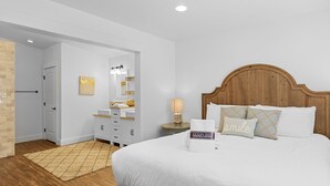 A bedroom with a wooden headboard, a bed with white linens, a nightstand with a lamp, a bathroom with a mirror and vanity, and a door leading to another room.