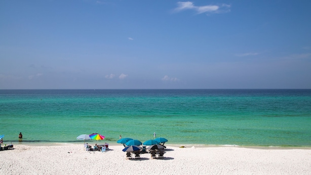 A beach with clear turquoise waters and a white sandy shore. There are several beach umbrellas and lounge chairs.