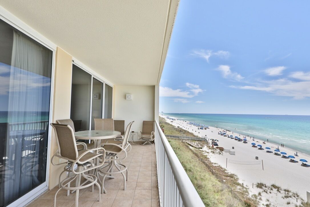 A balcony with a table and chairs overlooking a beach with umbrellas and people.