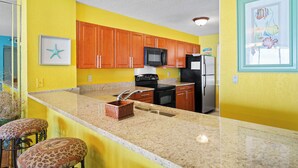 A kitchen with yellow walls, wooden cabinets, a black microwave, a refrigerator, and a granite countertop.