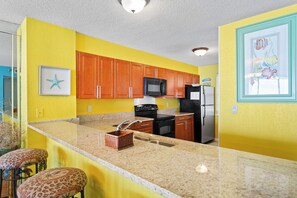 A kitchen with yellow walls, wooden cabinets, a black microwave, a refrigerator, and a granite countertop.