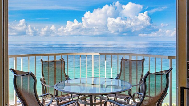 Varanda com mesa de vidro e quatro cadeiras, com vista para um mar azul e céu claro.