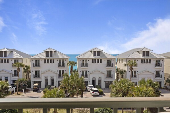 A row of white beachfront villas with balconies, parked cars, and palm trees.