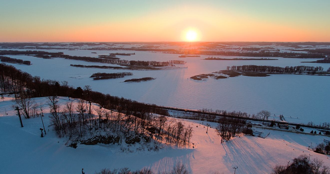 Paesaggio invernale con un lago ghiacciato, campi innevati e un ponte.