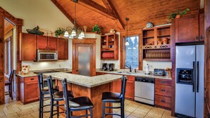 A kitchen with wooden cabinets, a central island with bar stools, a stainless steel refrigerator, and a window overlooking a forest. - On The Golf Course, Outdoor Kitchen, Hot Tub and So Much More! (Galena)