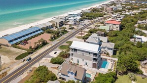 Vista aérea de uma área residencial costeira com uma casa imponente, tendo uma varanda grande e uma piscina.