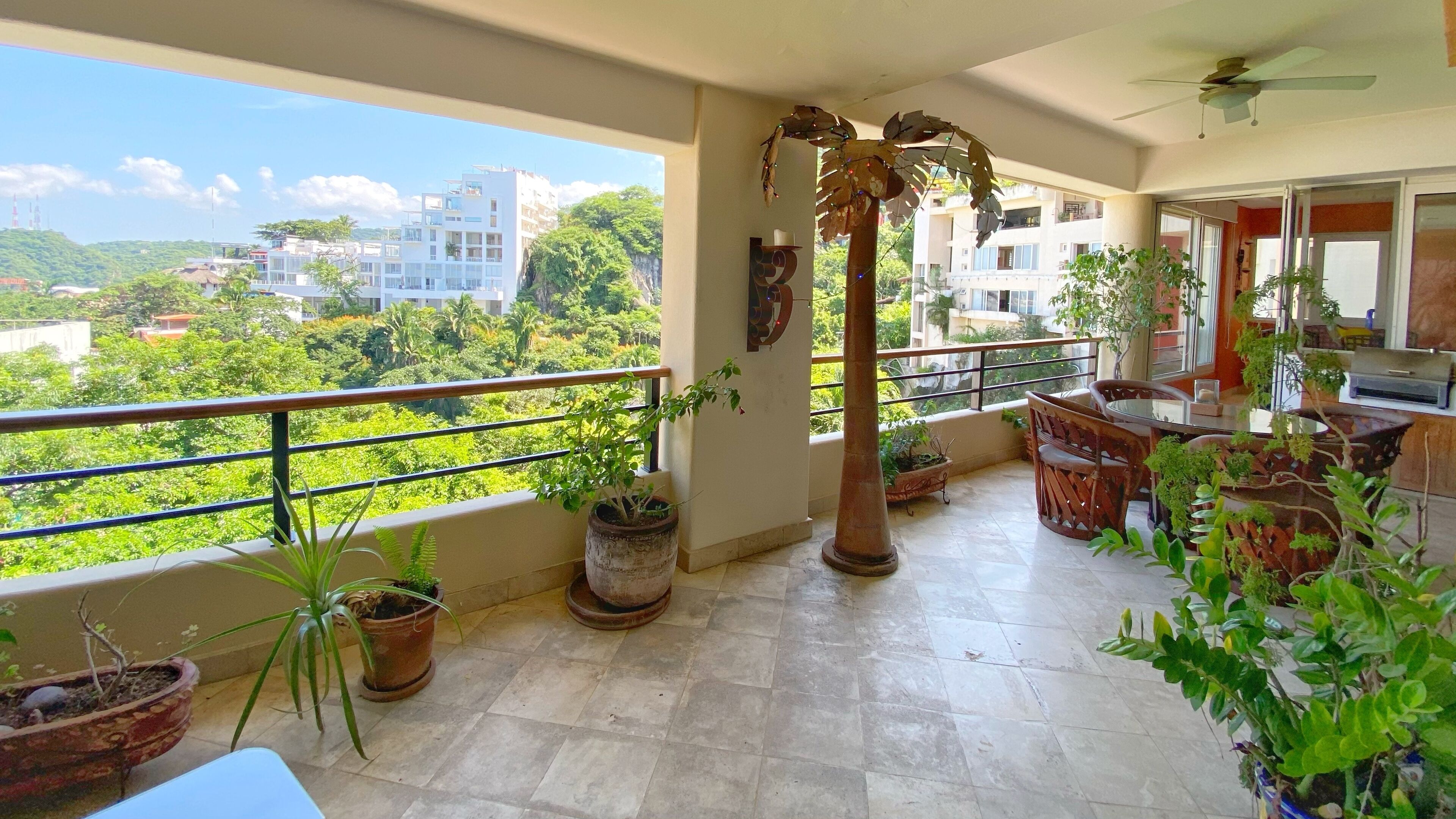 A balcony with potted plants, a ceiling fan, and a view of buildings and greenery.