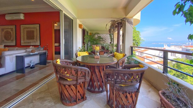 Un balcon avec une table ronde en verre, des chaises en osier et des coussins jaunes. On peut y admirer une vue sur la mer et les bâtiments en arrière-plan.