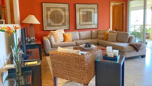Un salon avec un canapé beige, une table basse et un fauteuil en osier. Deux œuvres d’art encadrées ornent le mur, ainsi qu’une lampe et un vase rempli de fleurs, posé sur une table d’appoint.