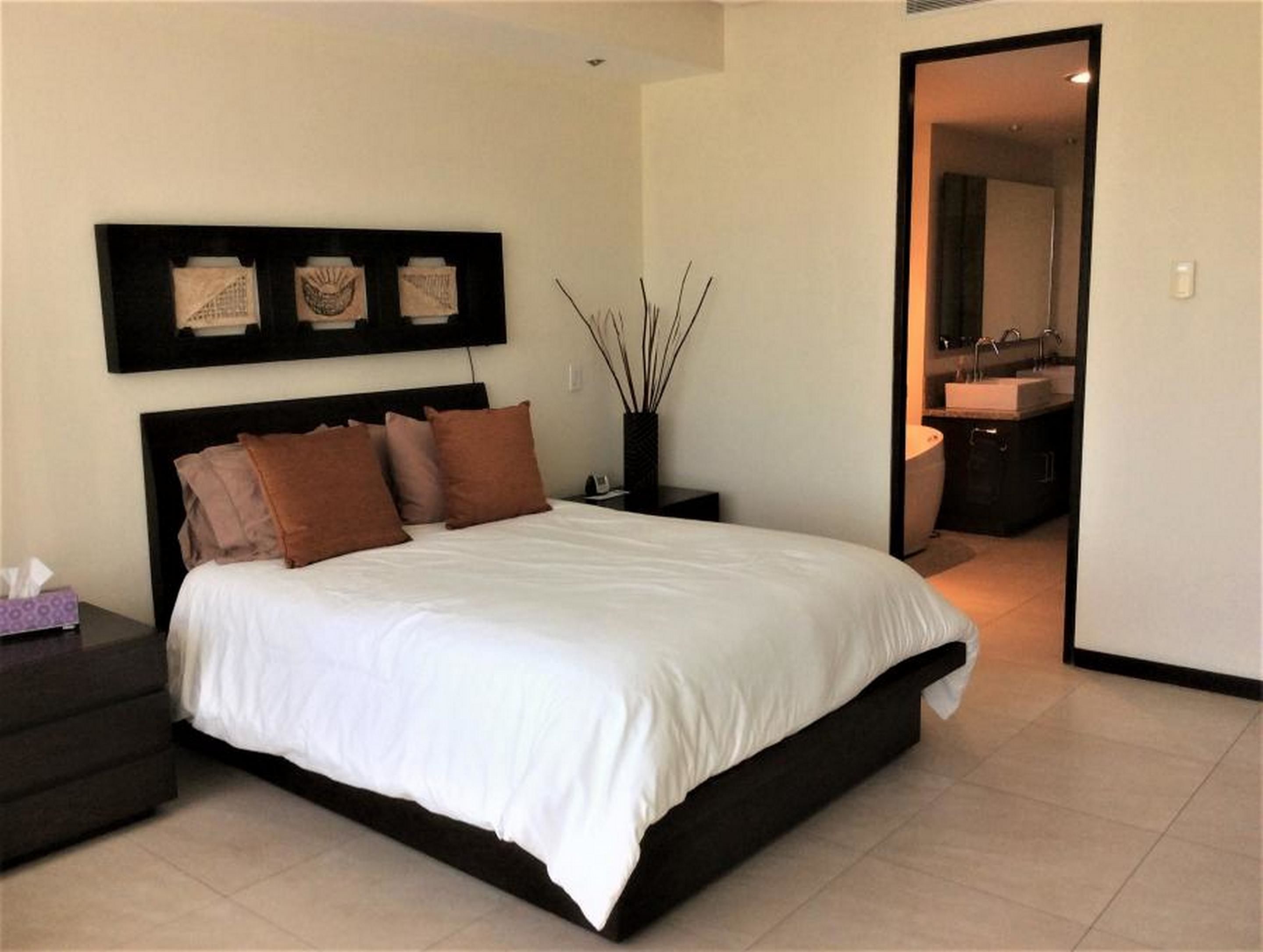 A modern bedroom with a large bed, a nightstand, a bedside table, a wall-mounted artwork, and a bathroom visible through an open door.