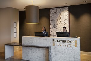 A reception desk at Staybridge Suites with a marble design, framed certificates on the wall, and a modern pendant light.