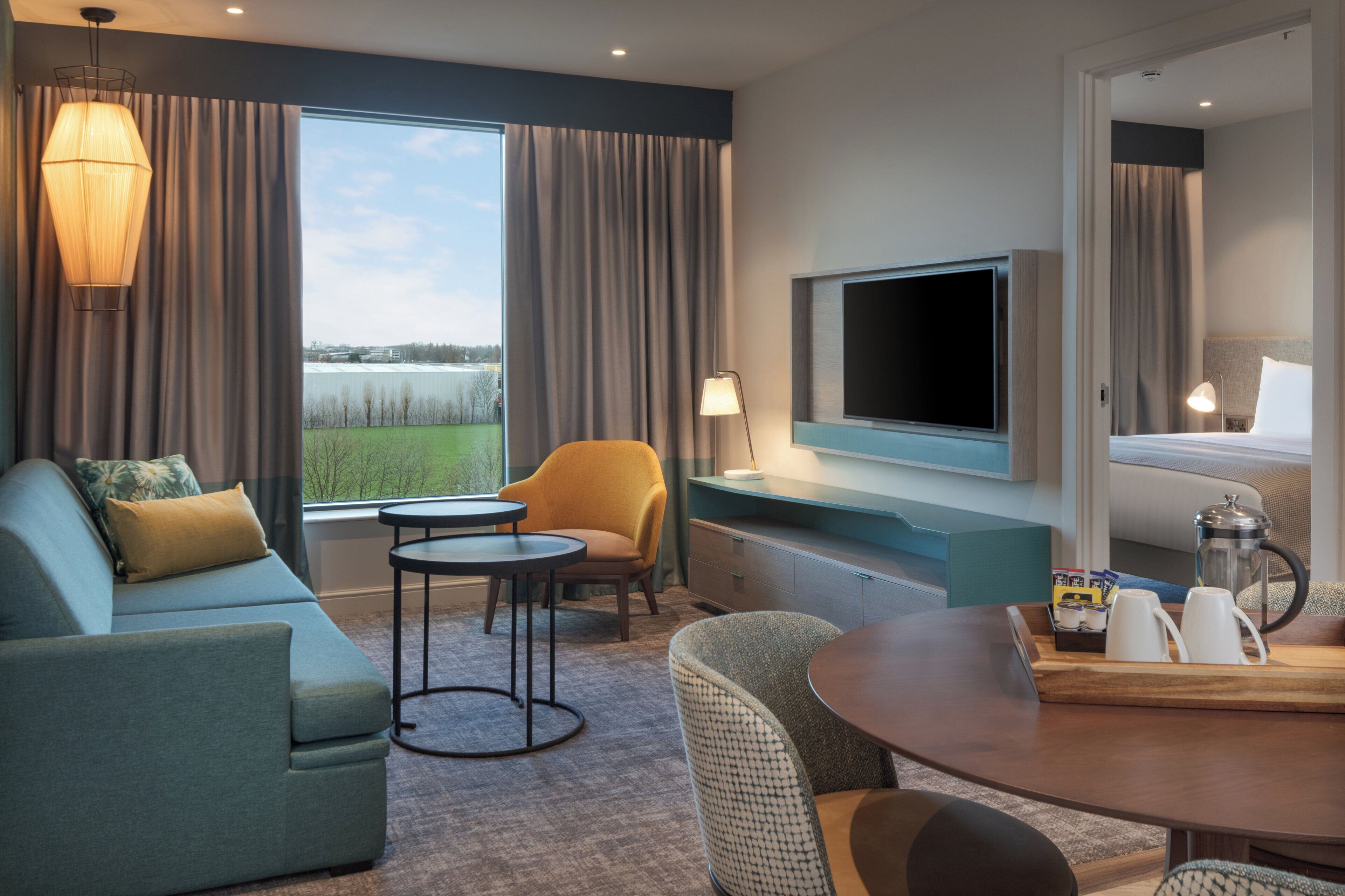 Ein Hotelzimmer mit Sofa, Ohrensessel, Esstisch, Fernseher und Blick ins Freie.