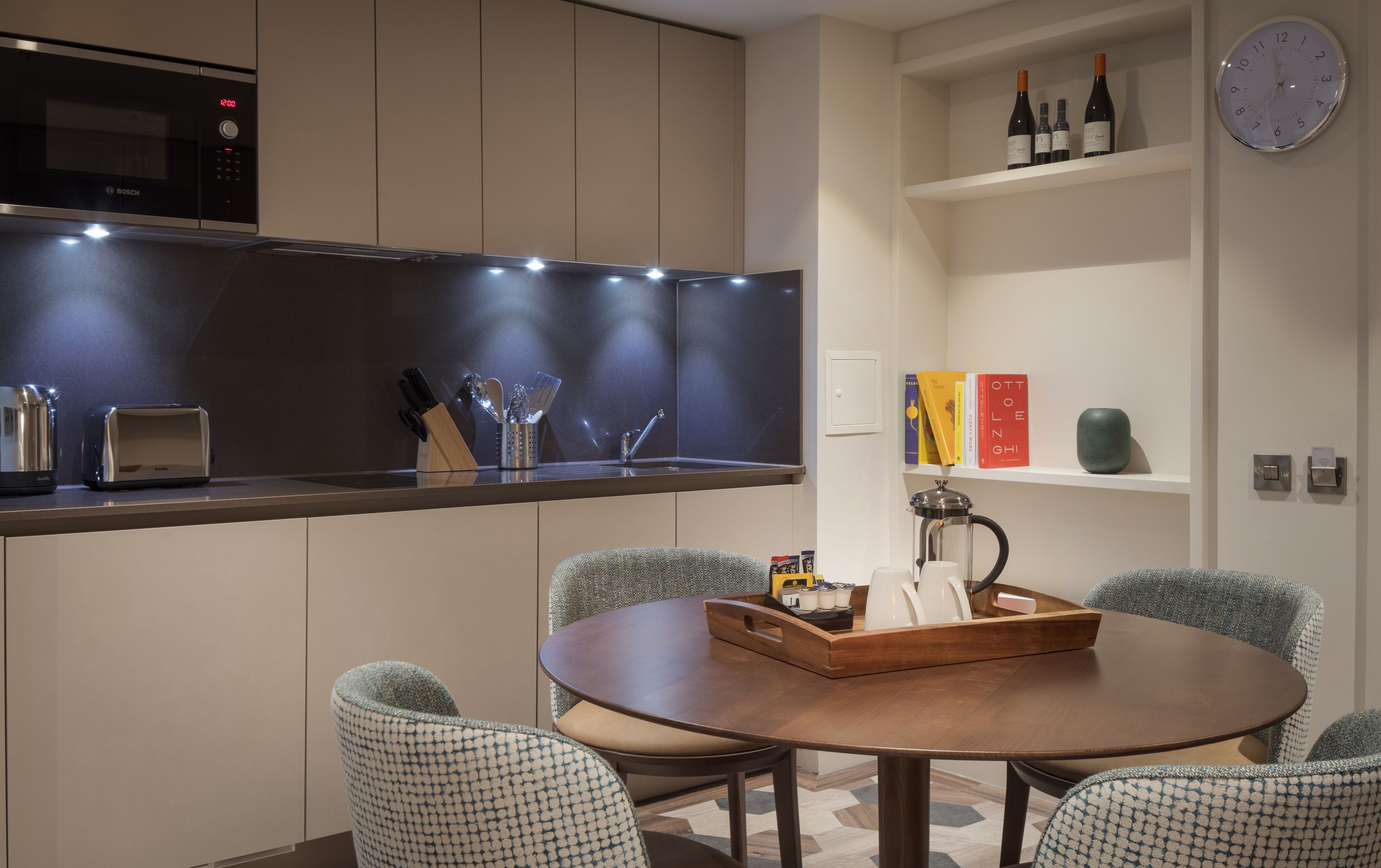 A modern kitchen with a Bosch microwave, a toaster, and a coffee maker. There is a dining table with chairs and a bookshelf with books and wine bottles.