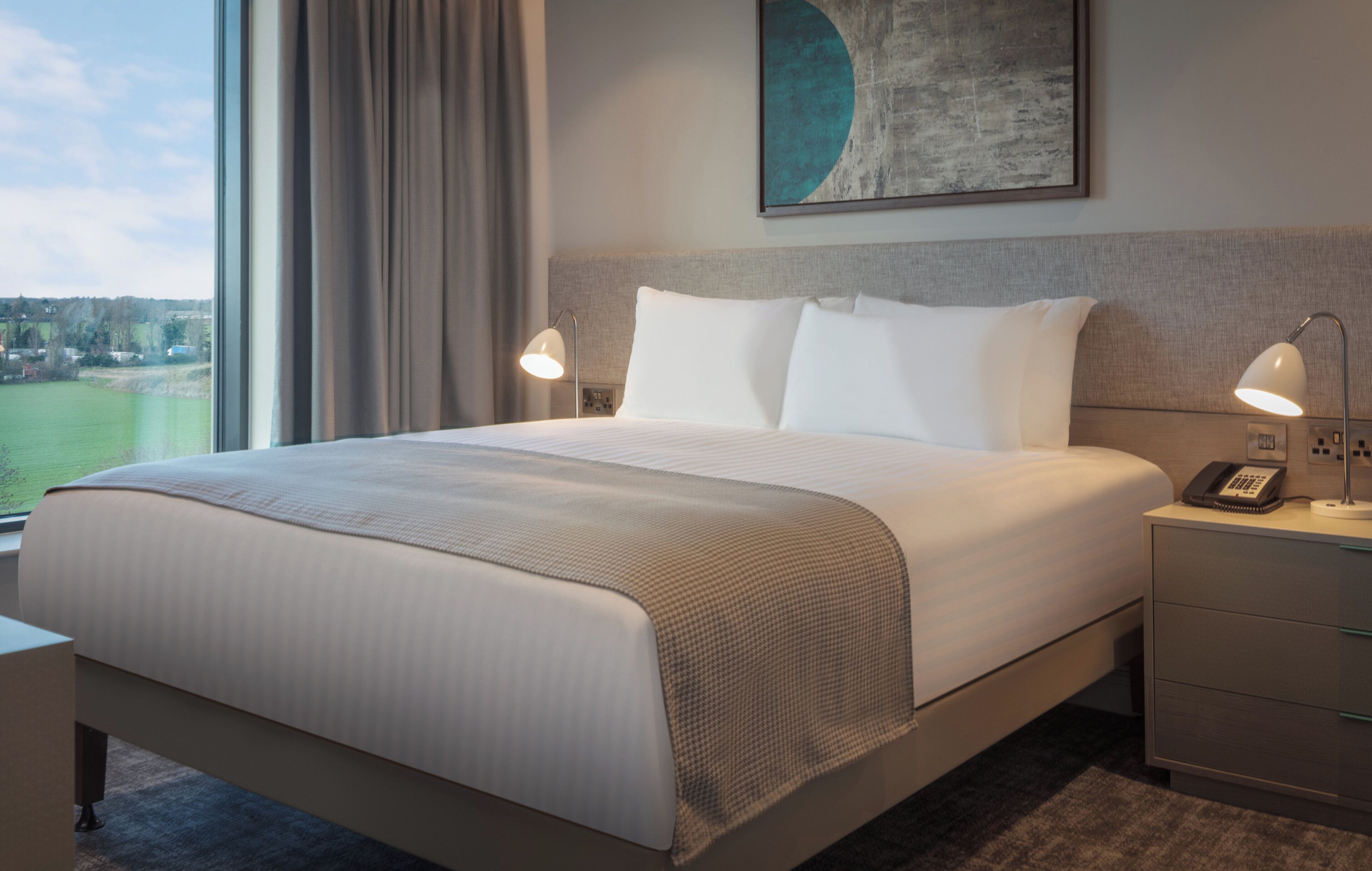 Quarto de hotel com uma cama grande, duas luminárias de cabeceira, um telefone e um quadro sobre a cama.