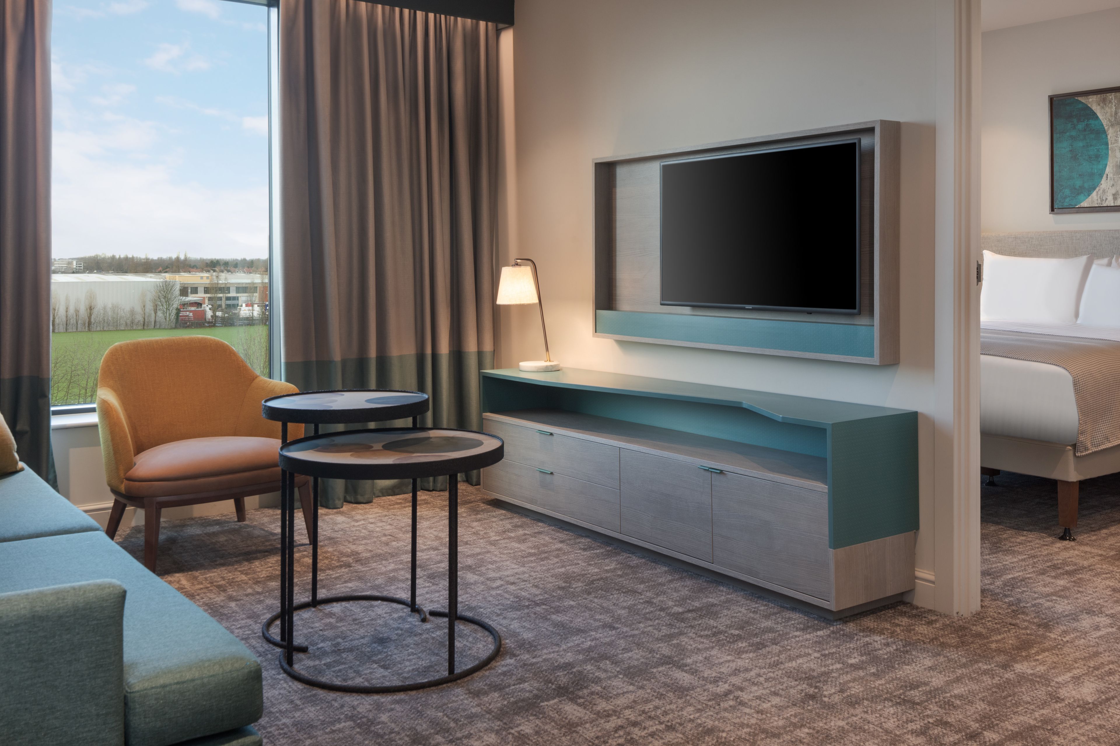 Habitación de hotel con una televisión de pantalla plana grande montada en la pared, un sofá, una silla, una mesita redonda y una cama con cabecero.
