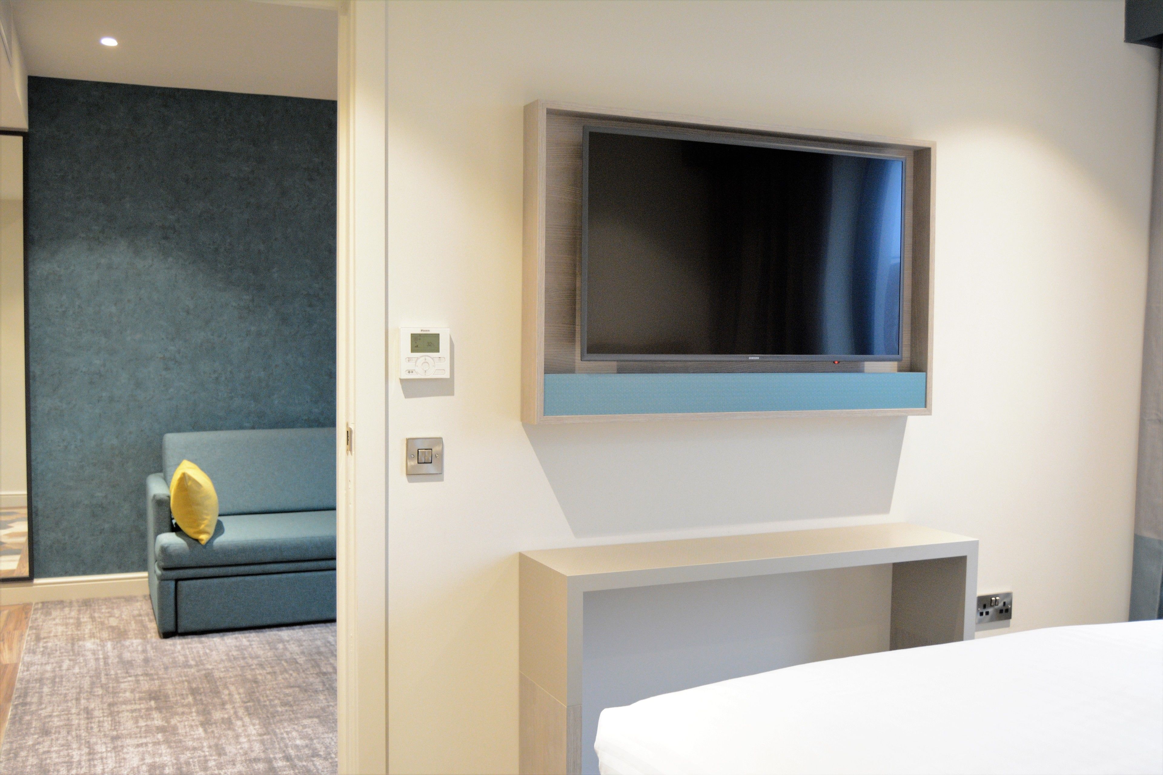 A modern hotel room with a large flat-screen TV mounted above a white console, a blue sofa with a yellow pillow, and a grey carpeted floor.
