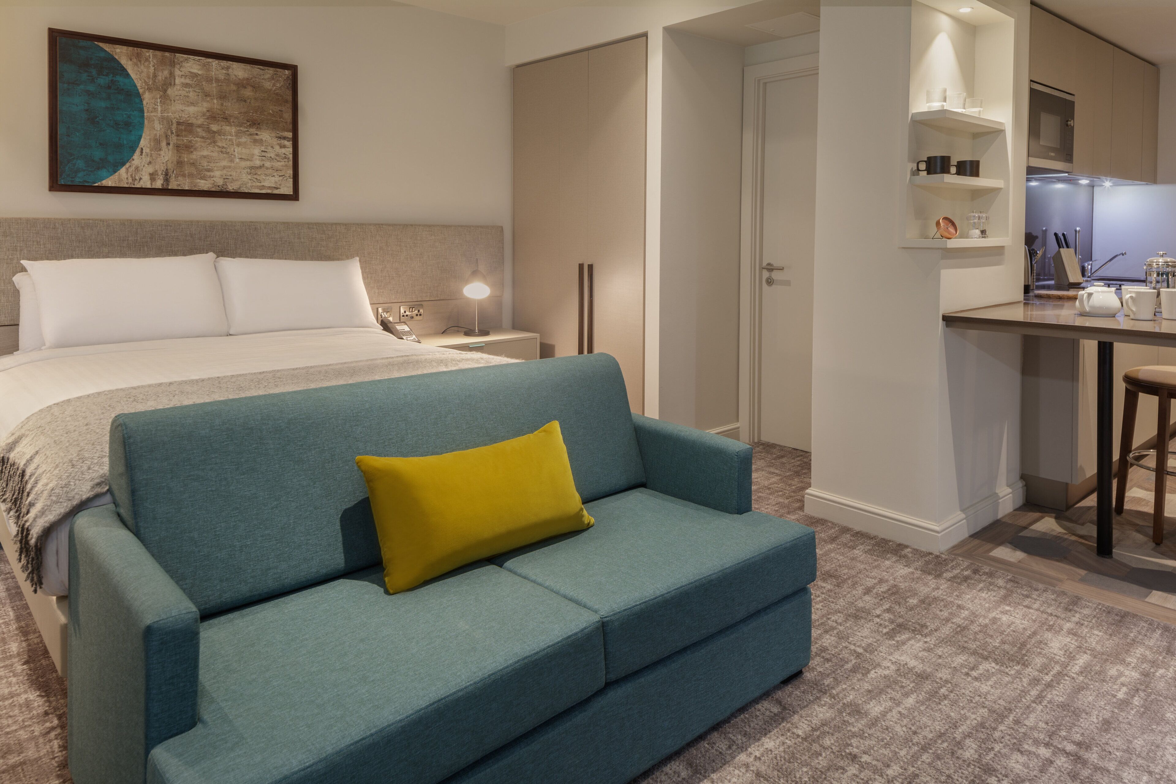 Een slaapkamer met een bed, een blauwe bank met een geel kussen, een nachtkastje met een lamp, en een keuken met een aanrecht en kasten.