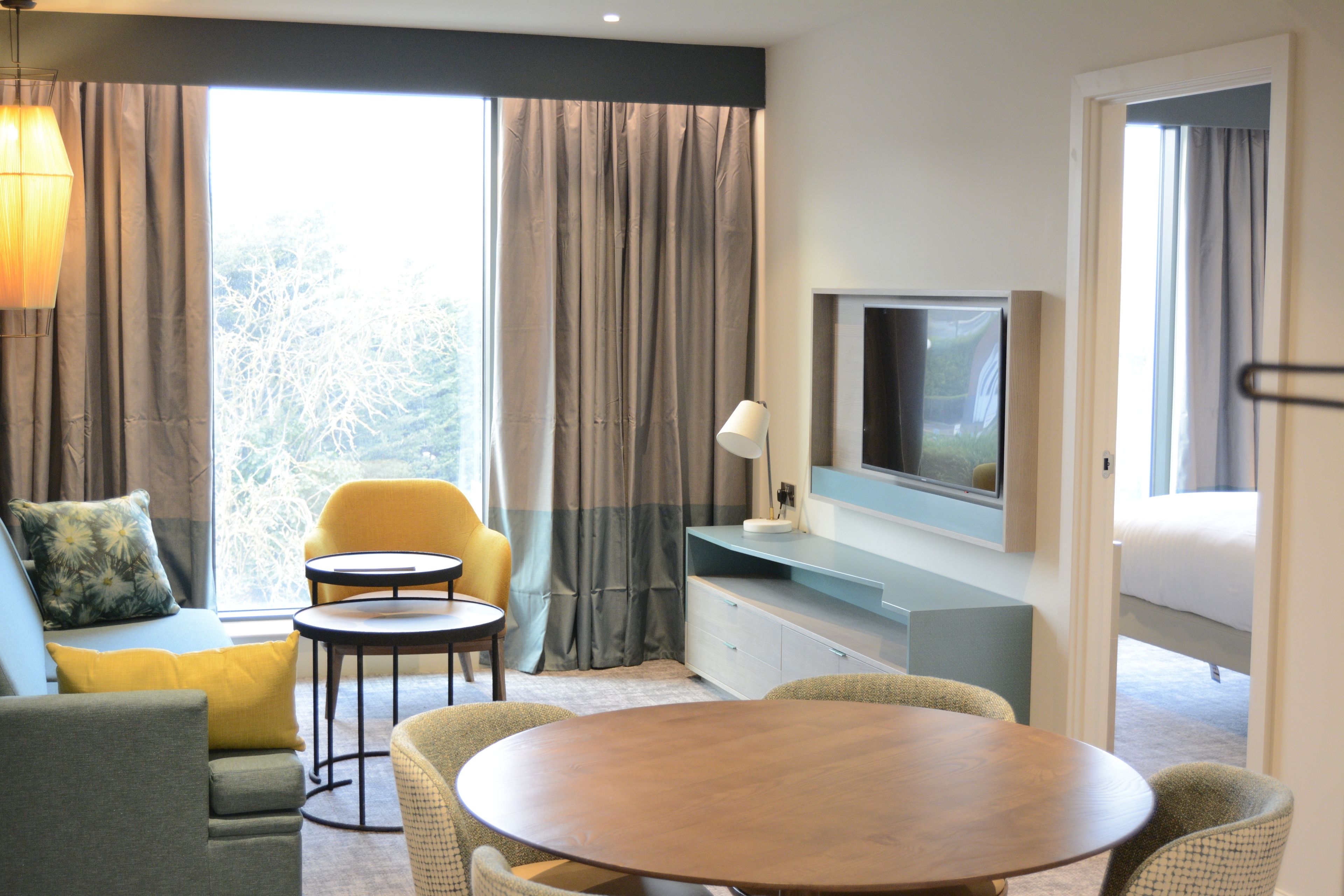 A modern hotel room with a sofa, yellow armchair, round dining table, TV, and a view of trees through the window.