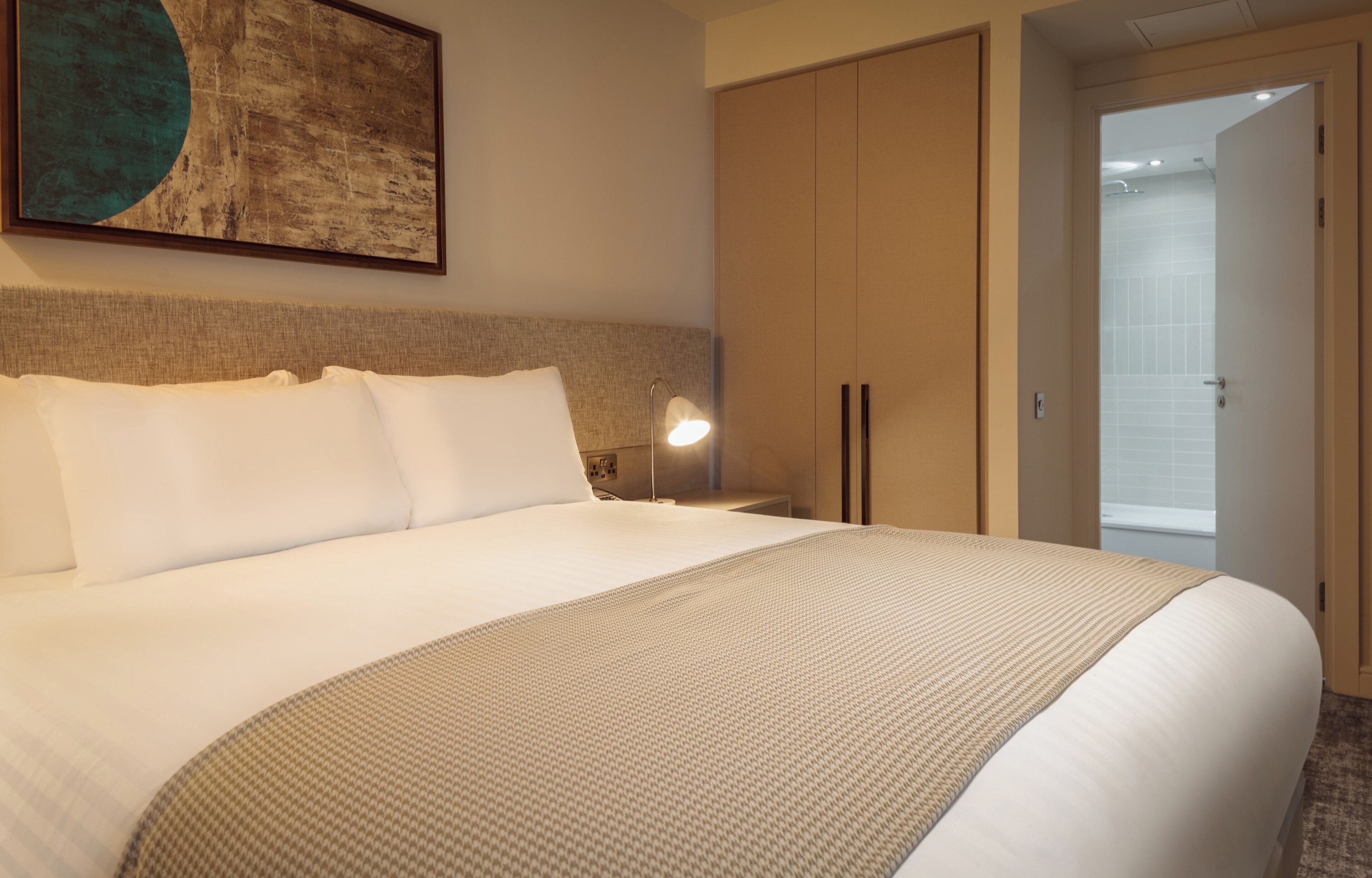 Habitación de hotel con una cama grande, una mesita de noche con lámpara, un armario y un baño visible a través de una puerta abierta.