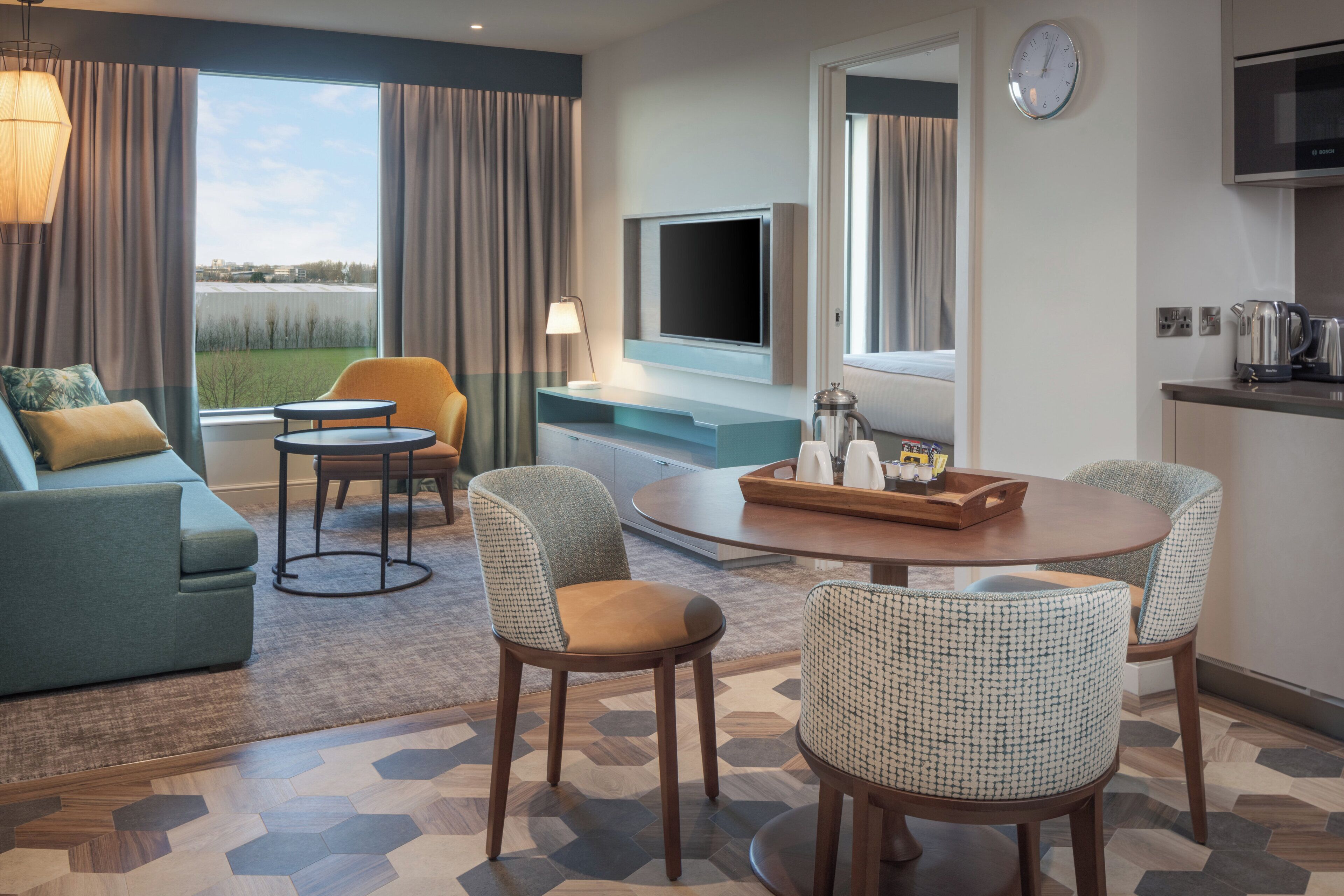 Quarto de hotel com sofá, cadeiras, TV, cafeteira e vista para o exterior.
