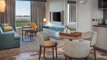 Quarto de hotel com sofá, cadeiras, TV, cafeteira e vista para o exterior.