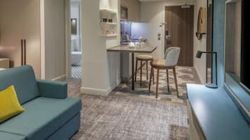 A modern living room with a blue sofa and a coffee table, a small kitchen area with a bar stool, and a view into the bathroom.