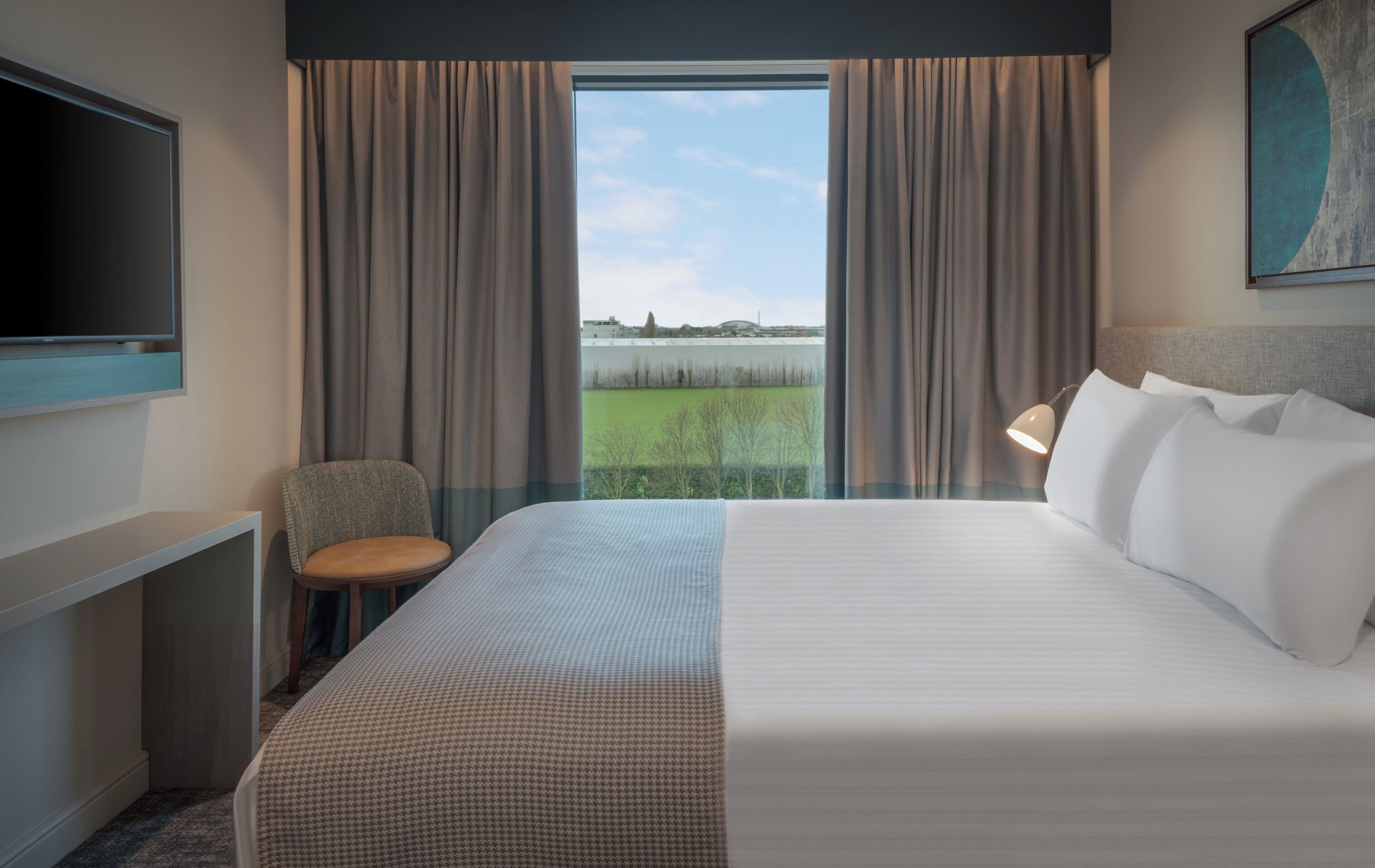 Ein Hotelzimmer mit Bett, Fernseher, Schreibtisch, Stuhl und Blick auf ein grünes Feld durch große Fenster.