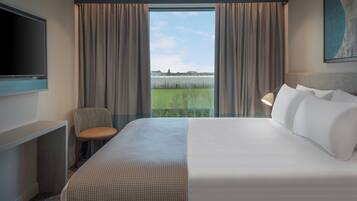 A hotel room with a bed, a TV, a desk, a chair, and a view of a green field through large windows.