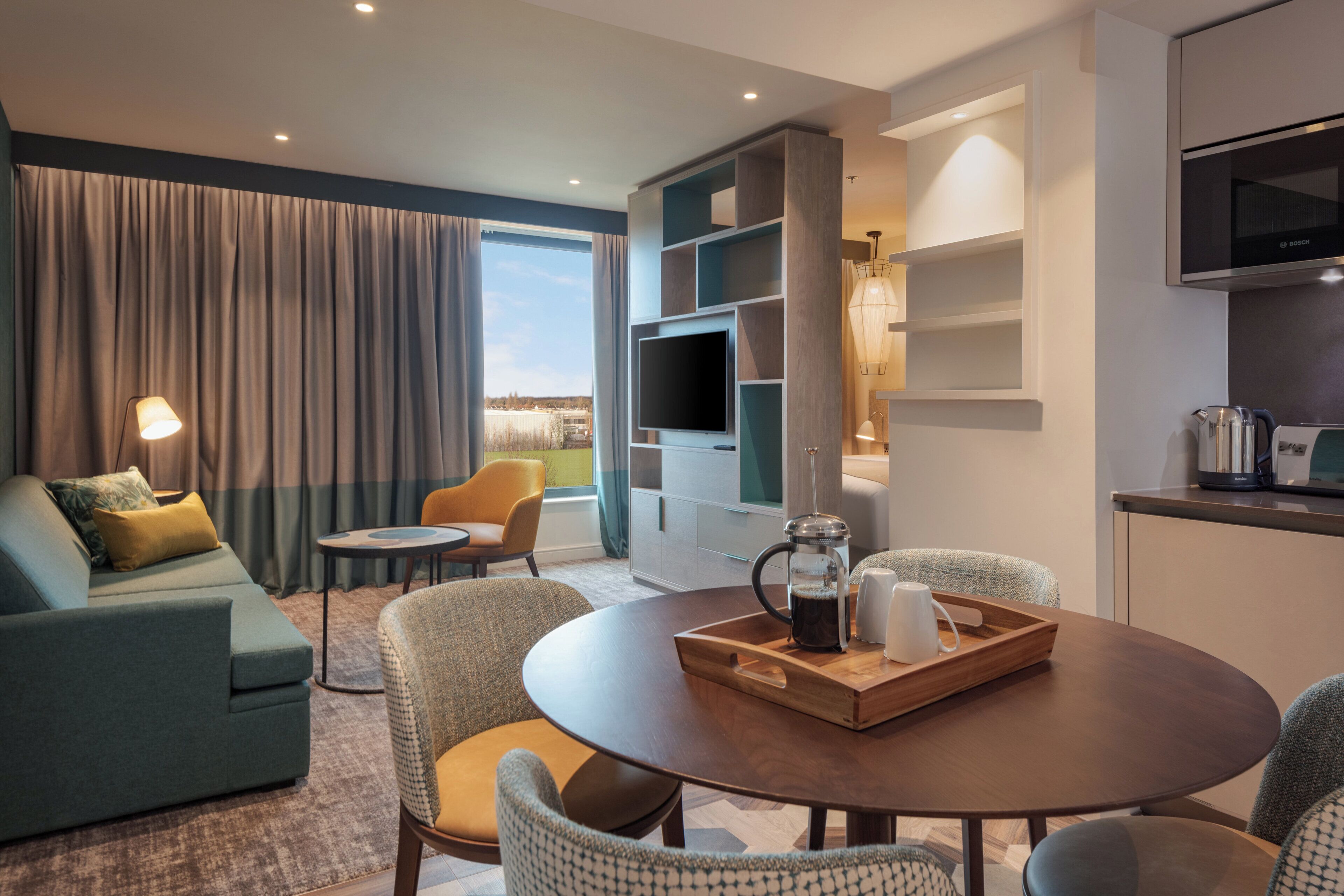 Une chambre d’hôtel moderne équipée d’un canapé, de fauteuils, d’une table à manger, d’un coin cuisine et d’une grande fenêtre avec des rideaux.
