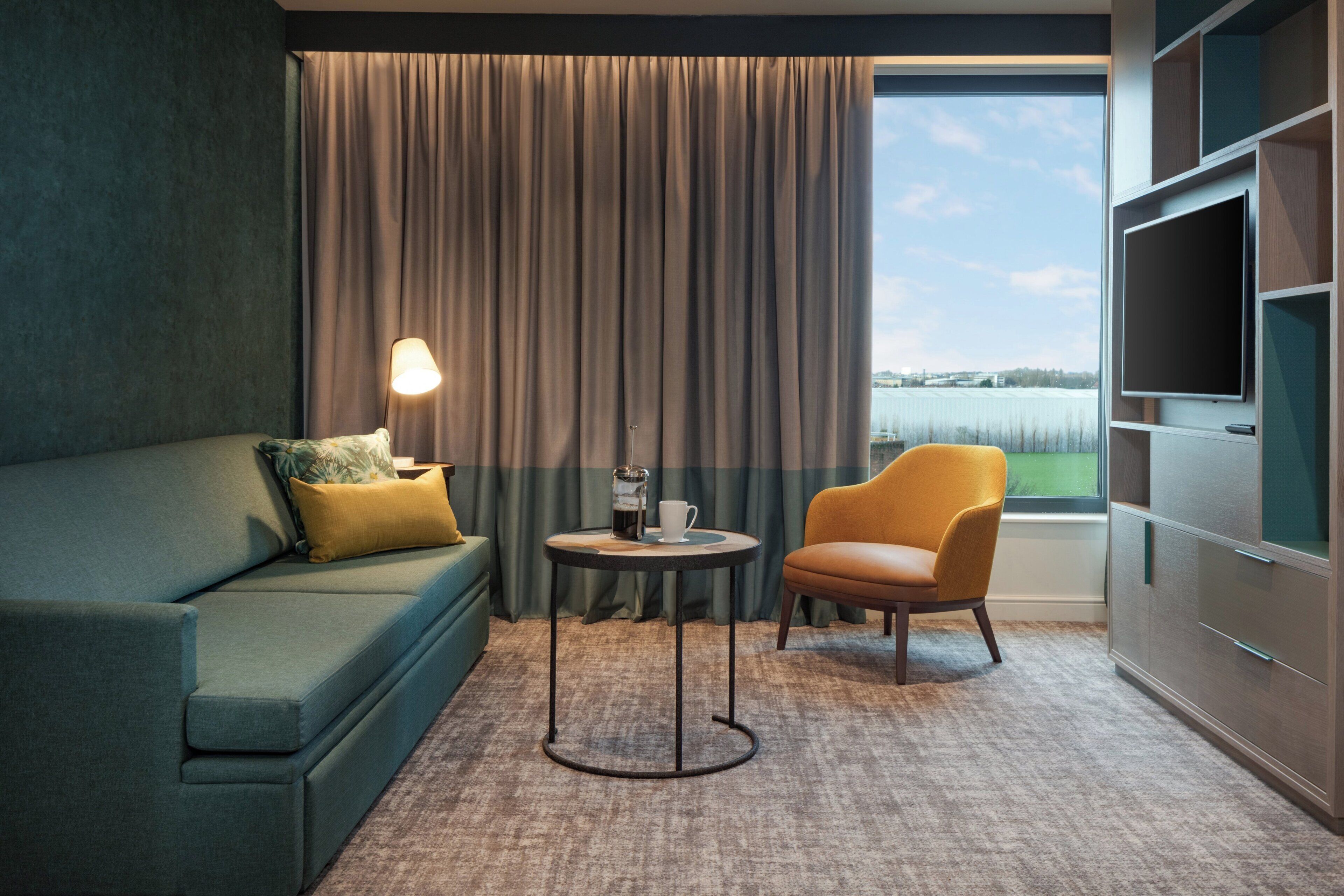 A hotel room with a sofa, armchair, coffee table, and a large window with curtains.