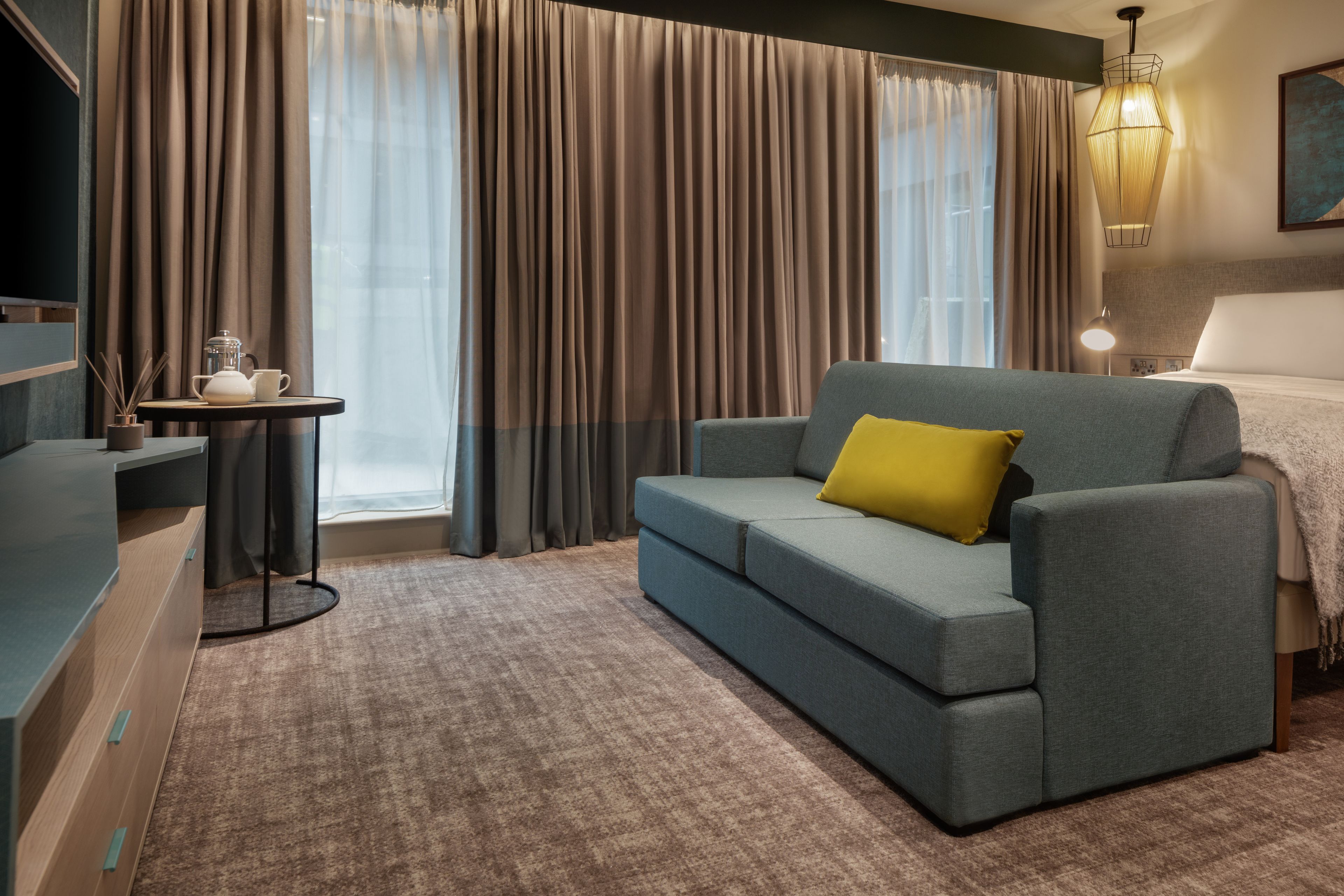 A modern hotel room with a grey sofa, a small table with a teapot, and a large window with curtains.