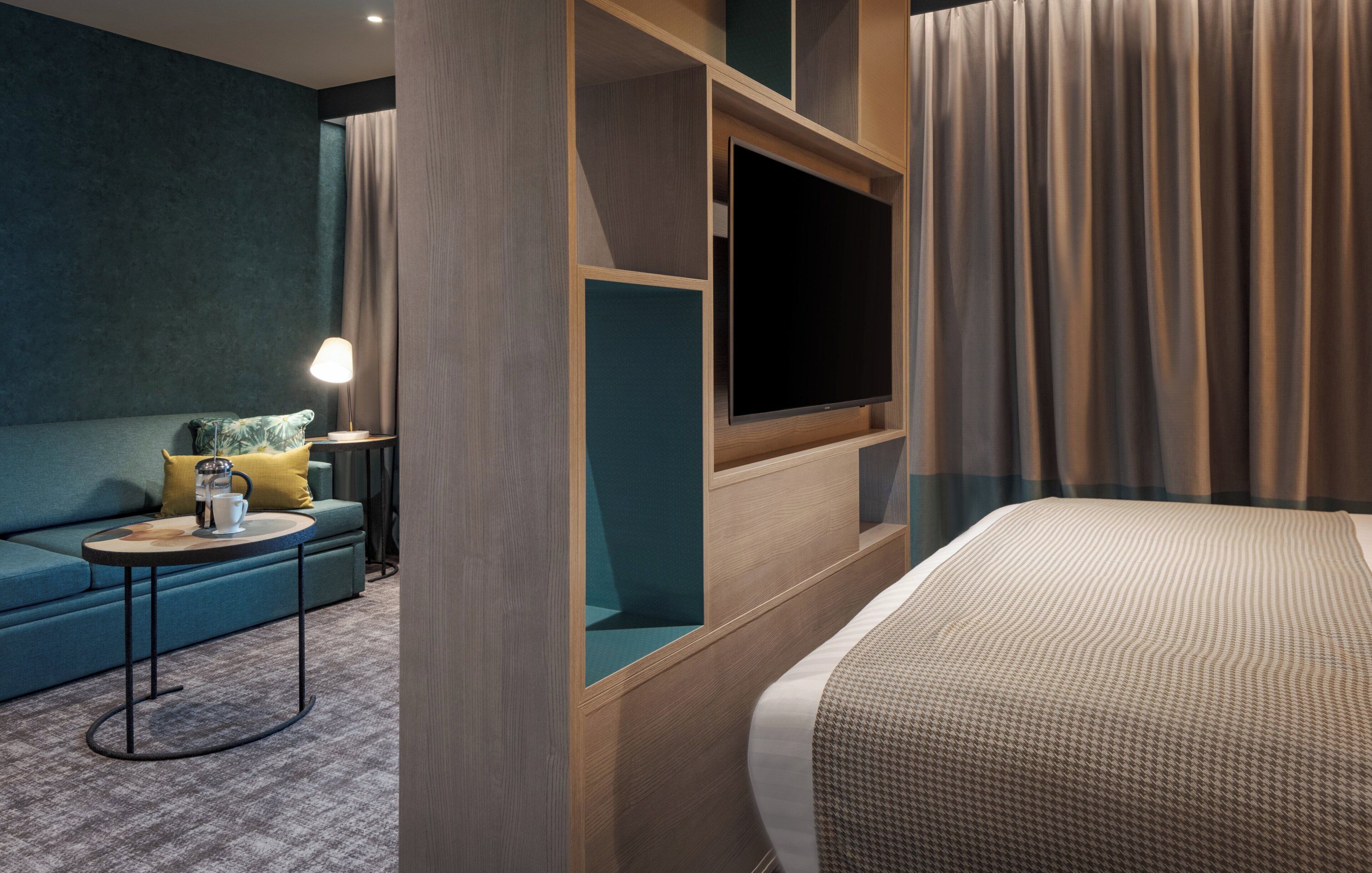 A modern hotel room with a bed, a sofa, a small table with a lamp, and a TV mounted on the wall.