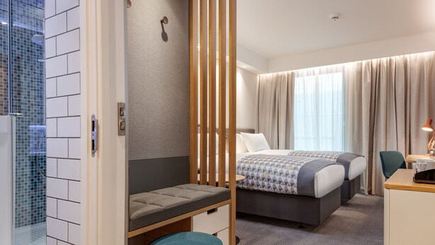 A hotel room with a bed, a desk, a chair, a small stool, a shower area, and a curtain. - Holiday Inn London Heathrow - Bath Road by IHG (West Drayton)