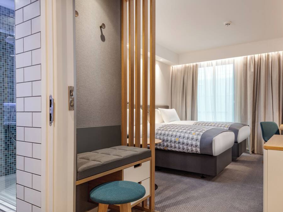 Habitación de hotel con cama, escritorio, una silla, un taburete pequeño, zona de ducha y ventana con cortinas.