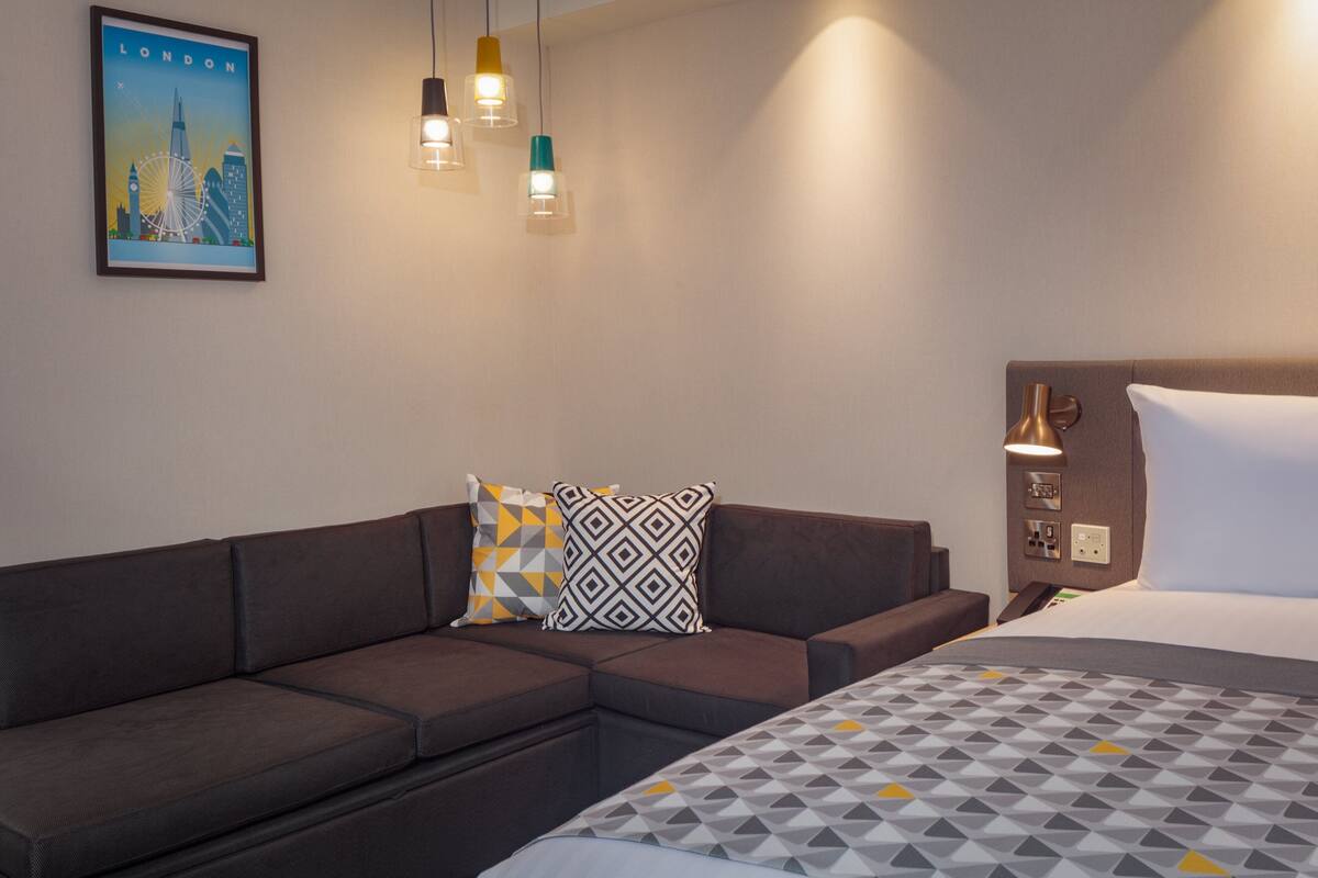 A modern hotel room with a sectional sofa, a bed with a geometric-patterned duvet, a cityscape poster on the wall, and pendant lighting.