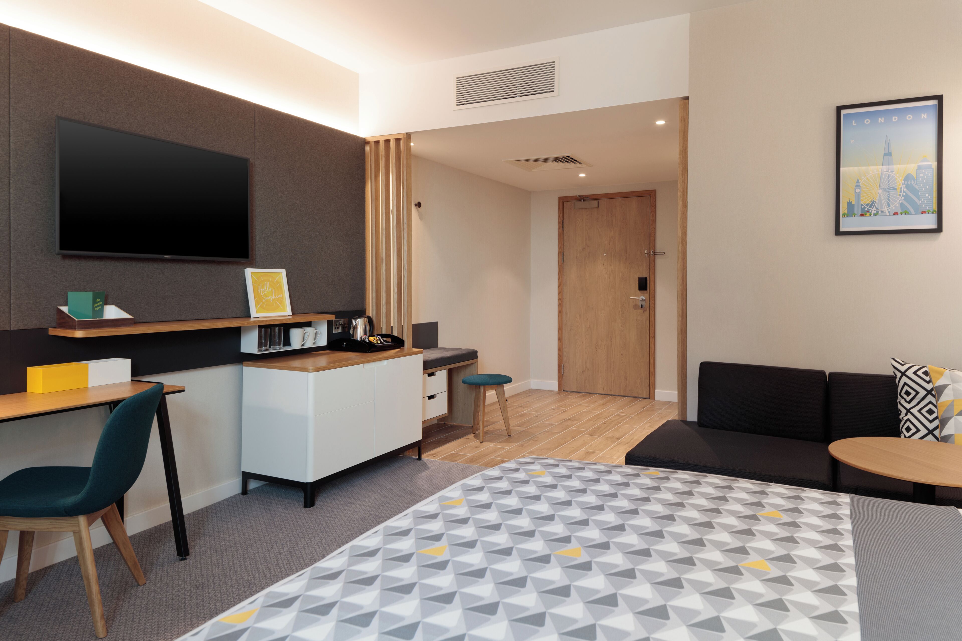 A hotel room with a bed, a desk, a chair, a sofa, a TV, a picture frame, and a door leading to another room.