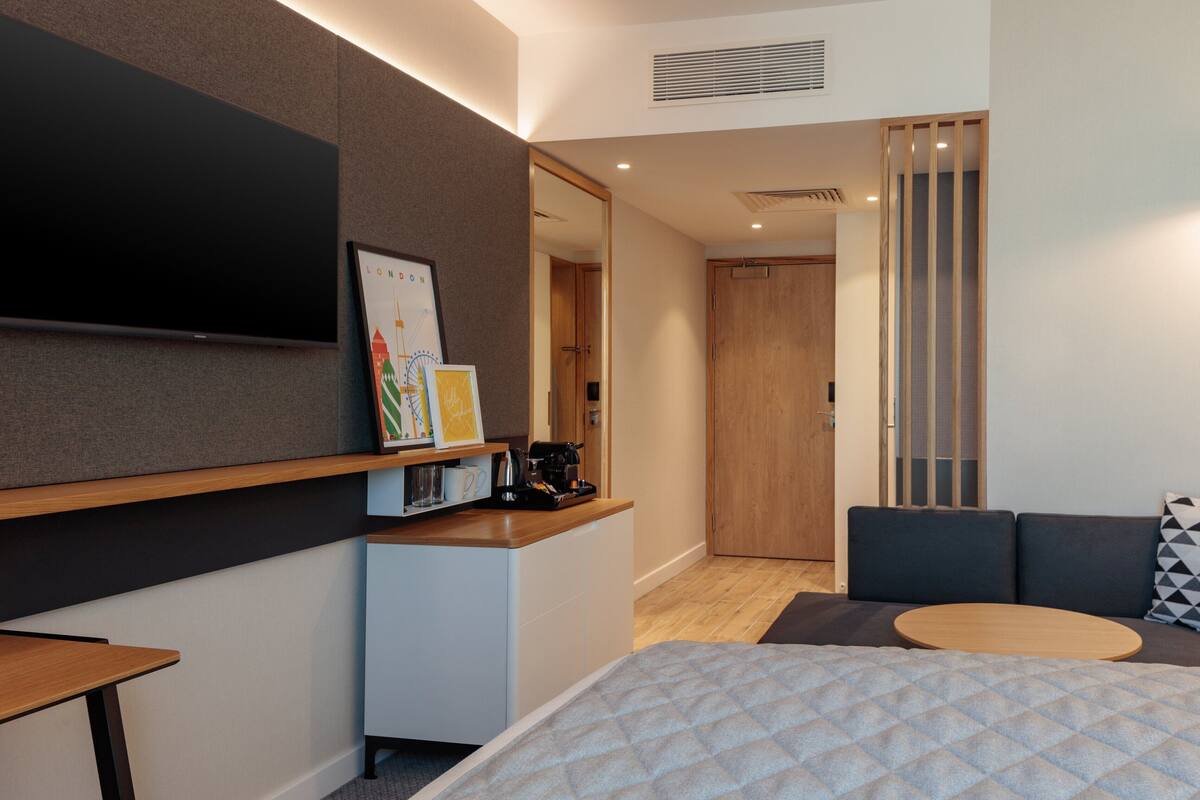 A modern hotel room with a bed, a small desk, a sofa, and a TV mounted on the wall.