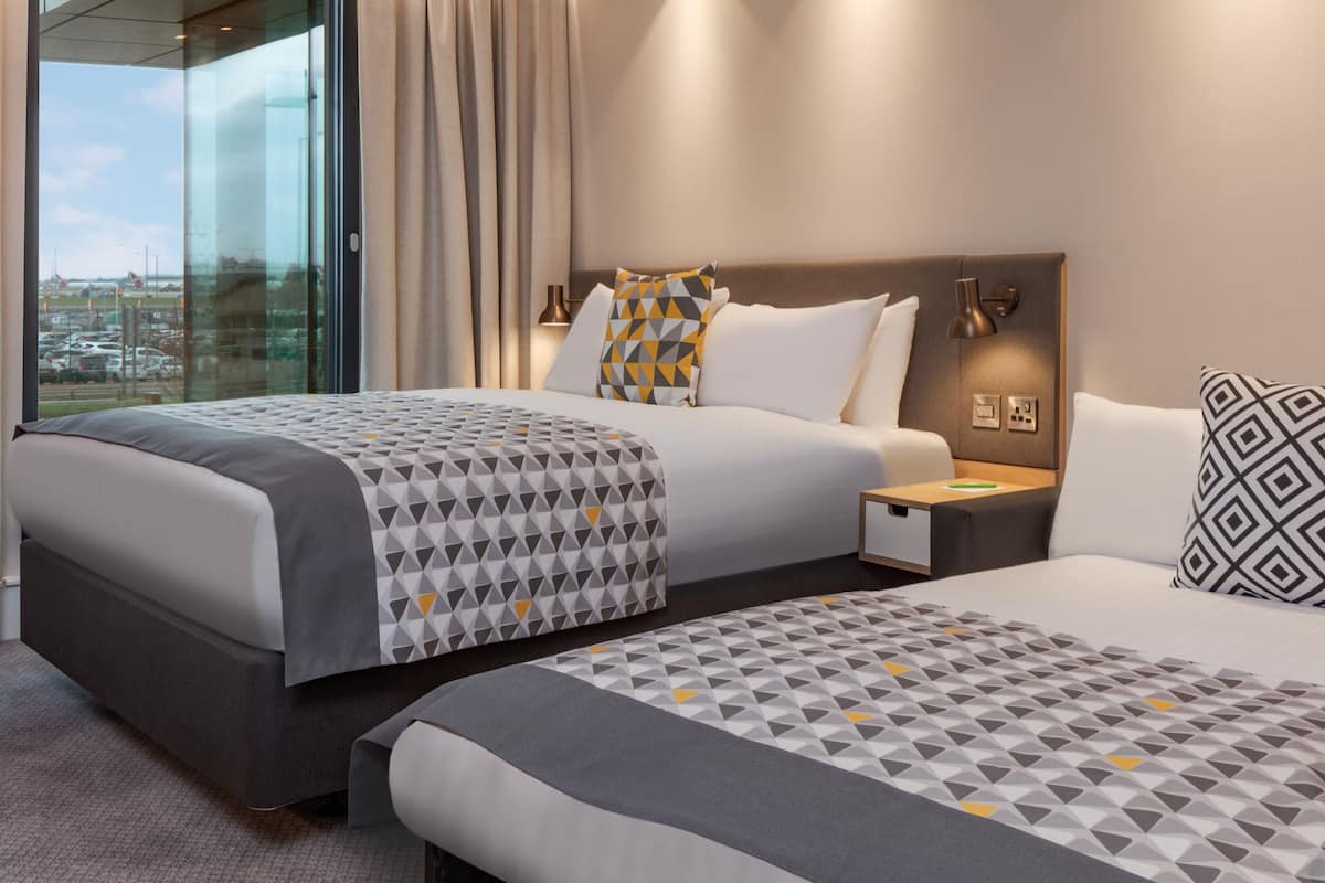 A hotel room with two beds, a large window with curtains, a nightstand, and a lamp.