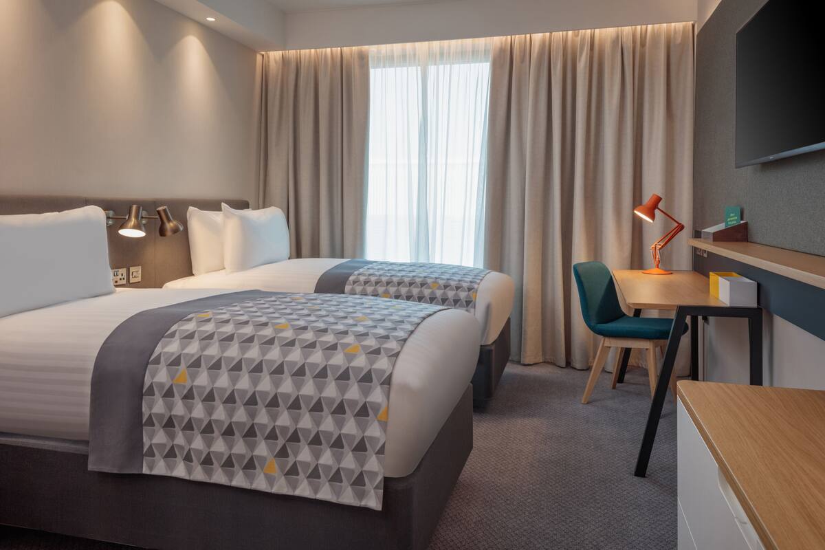 A hotel room with two beds, a desk, a chair, a TV, and a window with curtains.