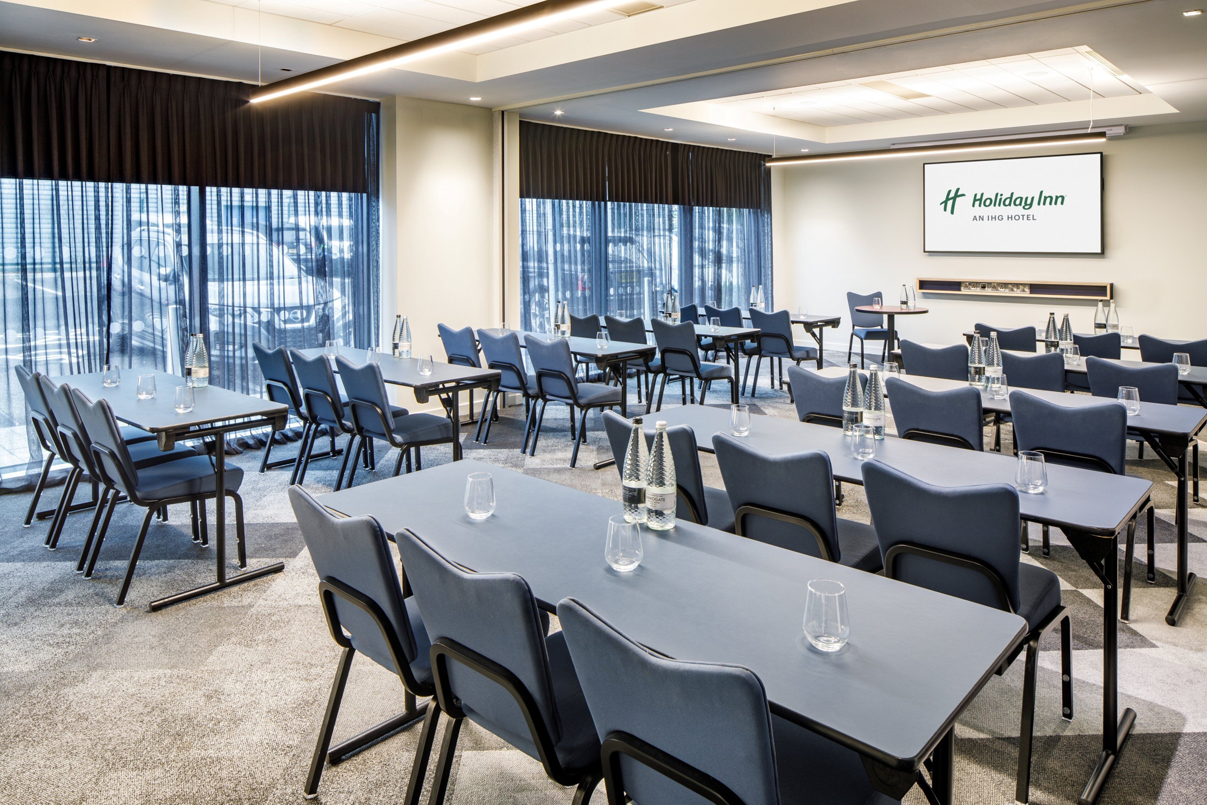 Un salón de conferencias con mesas y sillas, una pantalla grande mostrando el logotipo de Holiday Inn y amplias ventanas con cortinas.