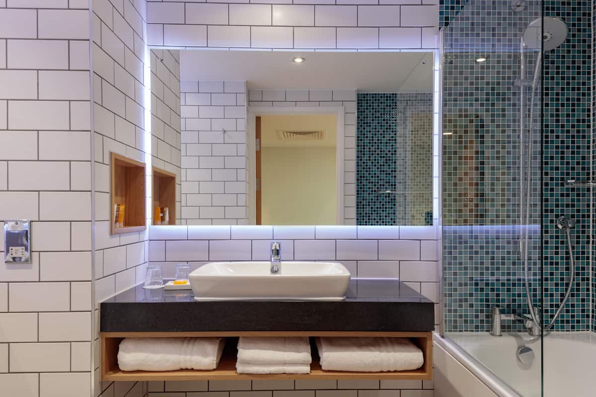Un baño moderno con pared de azulejos blancos, encimera negra, lavamanos blanco y zona de ducha con mampara de vidrio.